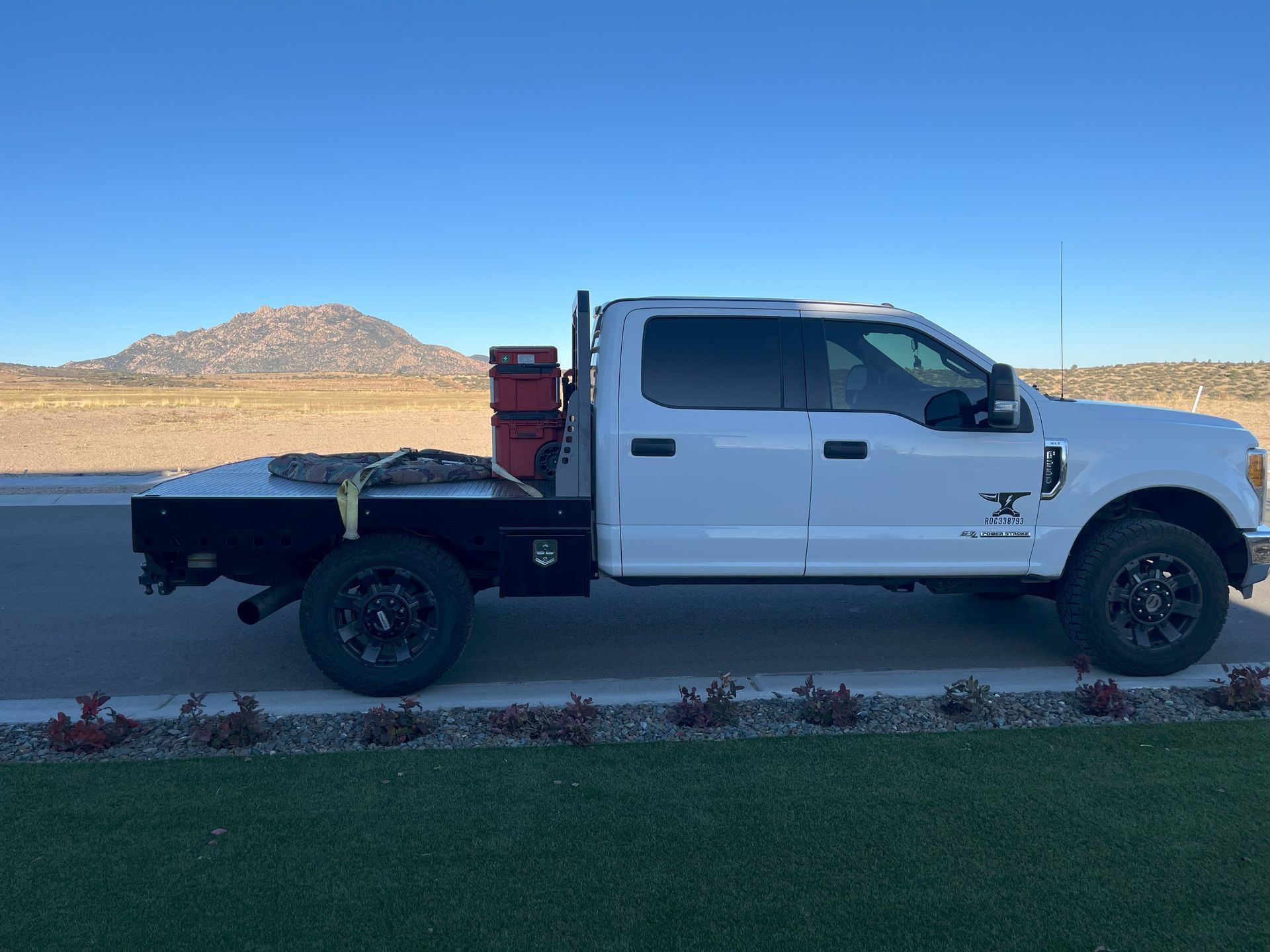 White flatbed truck parked on asphalt, green grass in front, dry landscape background.