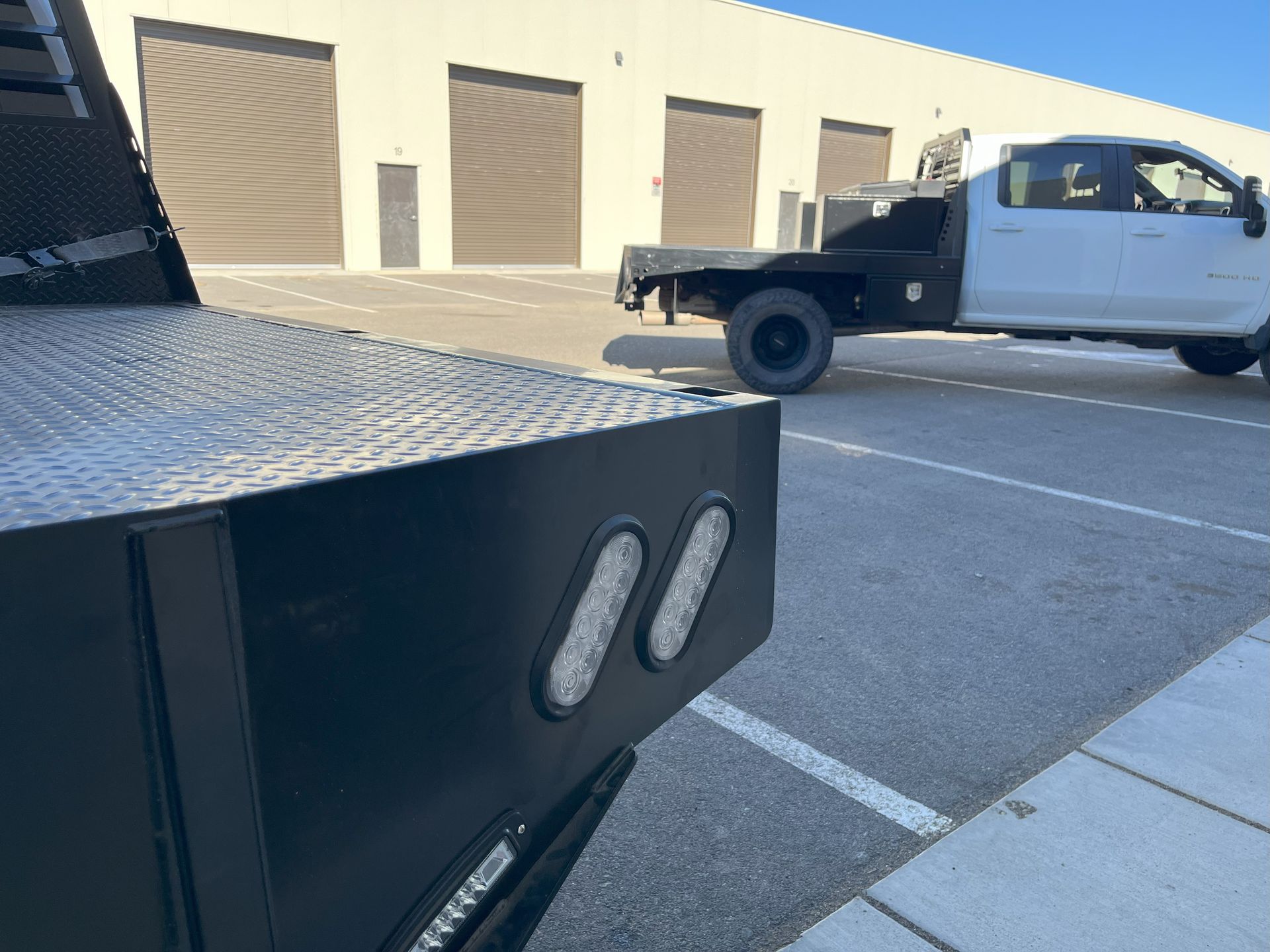 Black flatbed truck with LED lights, parked in front of a white truck and a warehouse.