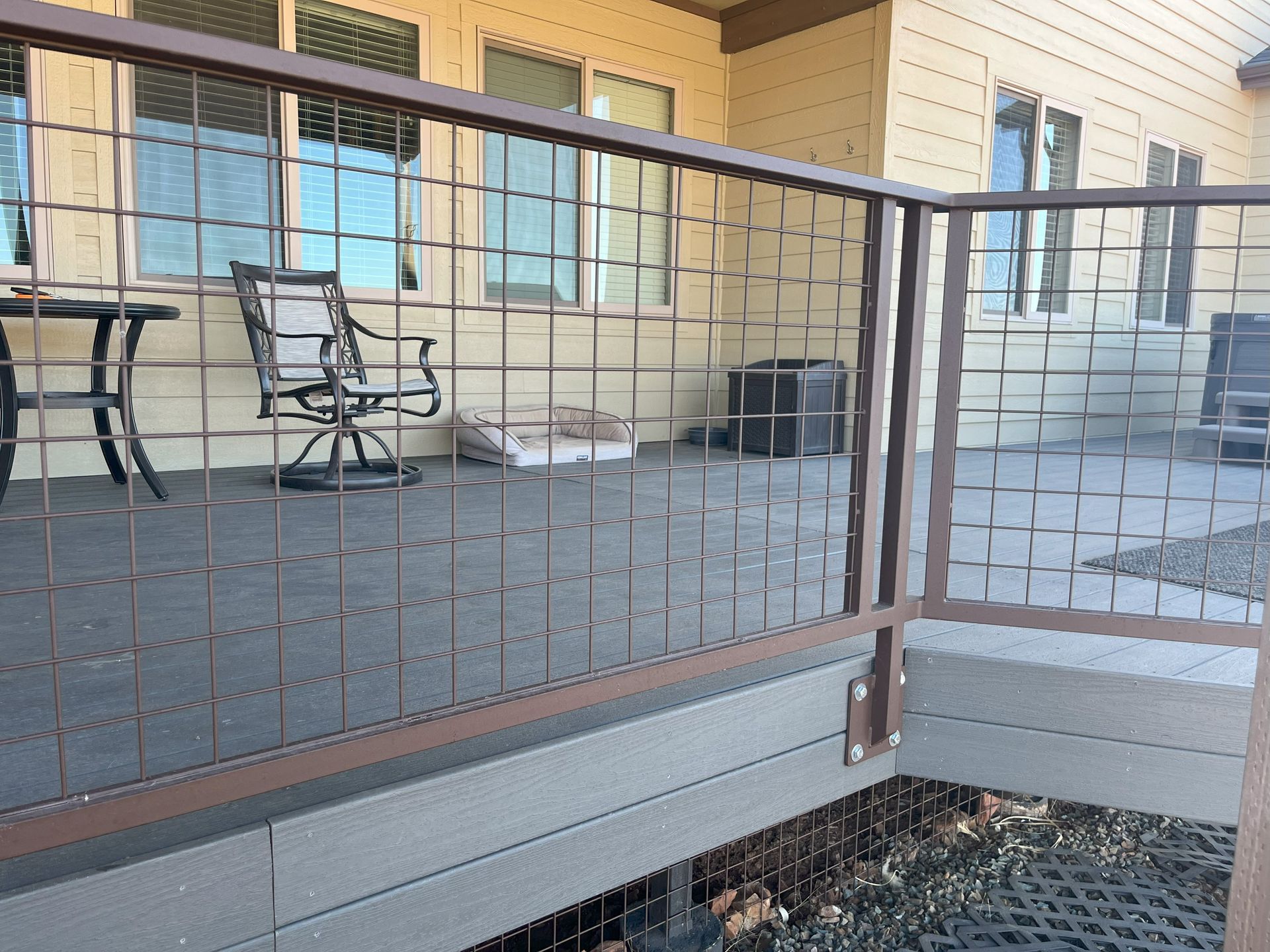 Deck with metal mesh railing and brown framing, looking out to windows and furniture.