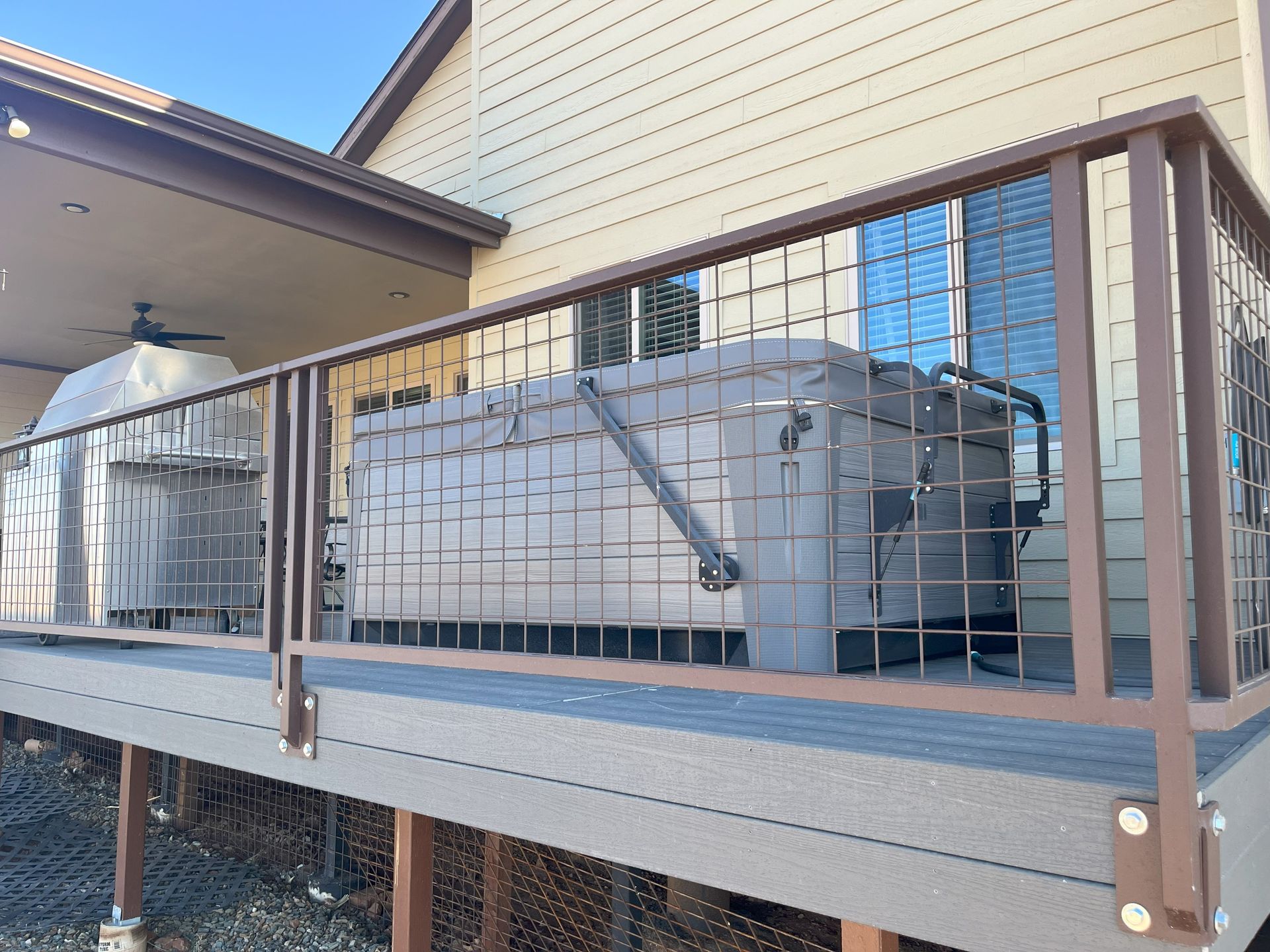 Hot tub on a deck with brown railing, adjacent to a house with beige siding. Covered grill nearby.