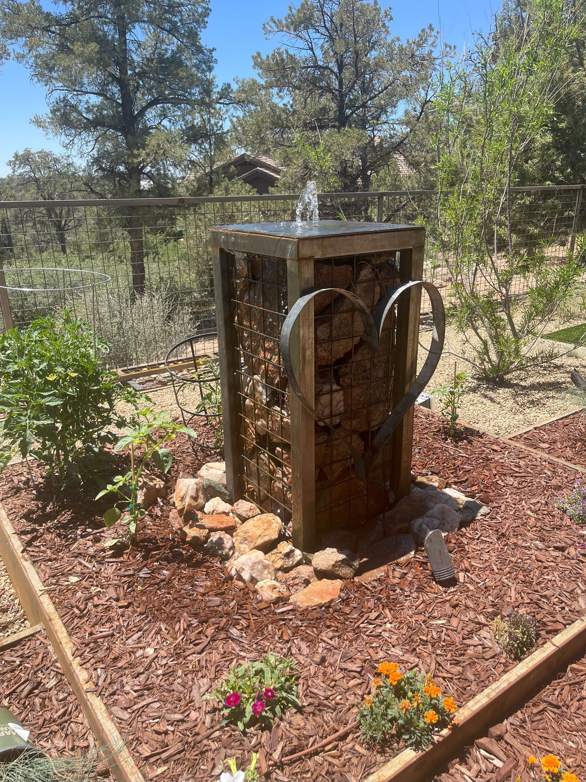 Outdoor gabion fountain with flowing water, surrounded by rocks and garden.