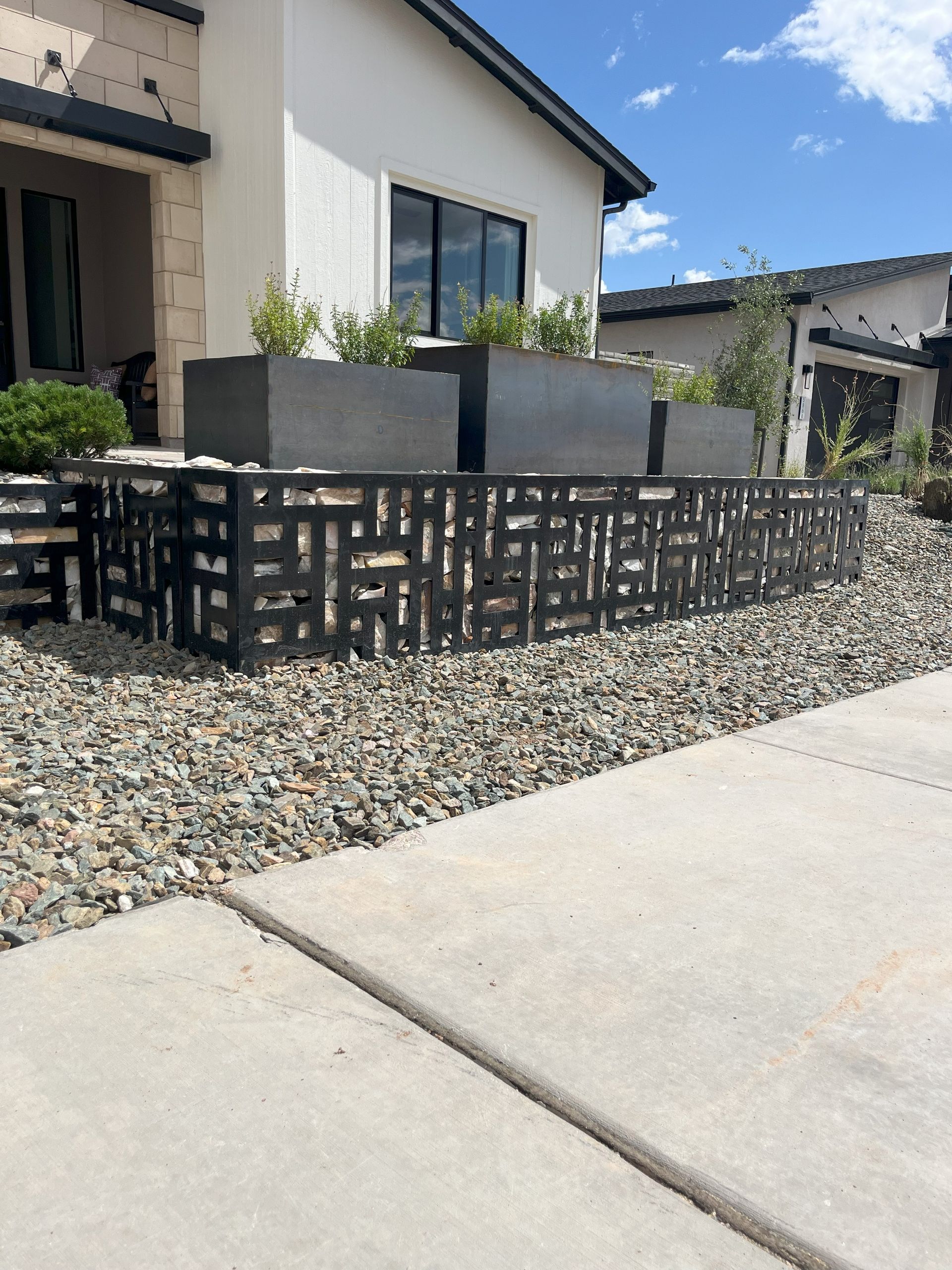 Black metal fence with decorative cutouts in front of a modern home with planters and gravel.