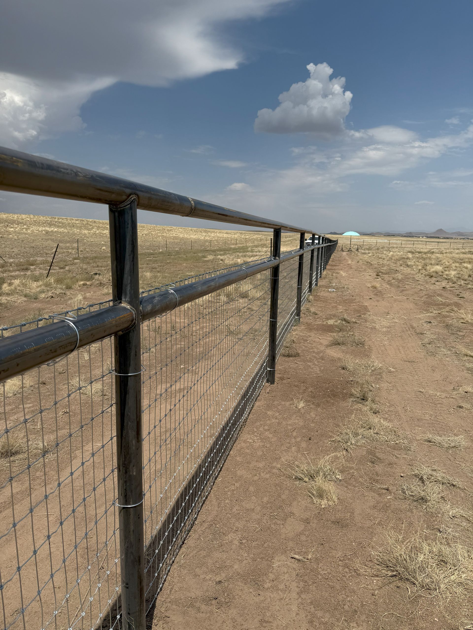 Metal fence stretches across a brown field under a blue sky with clouds.
