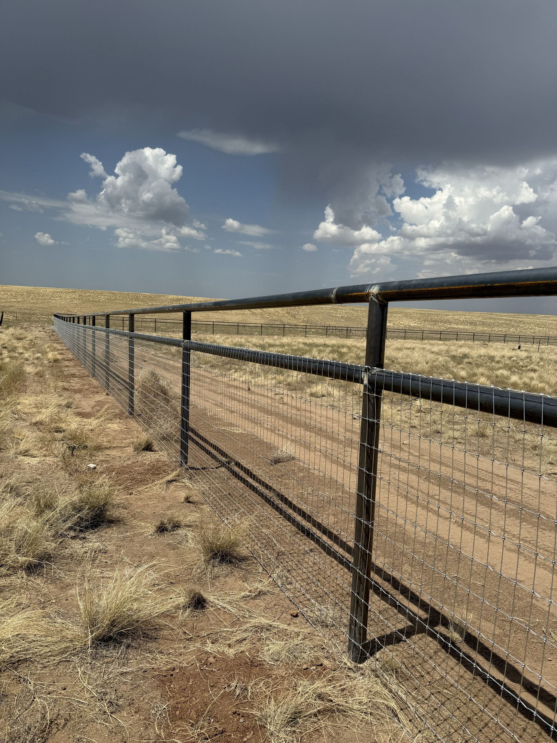 A black pipe fence with wire mesh runs through a dry, grassy field under a cloudy, overcast sky.