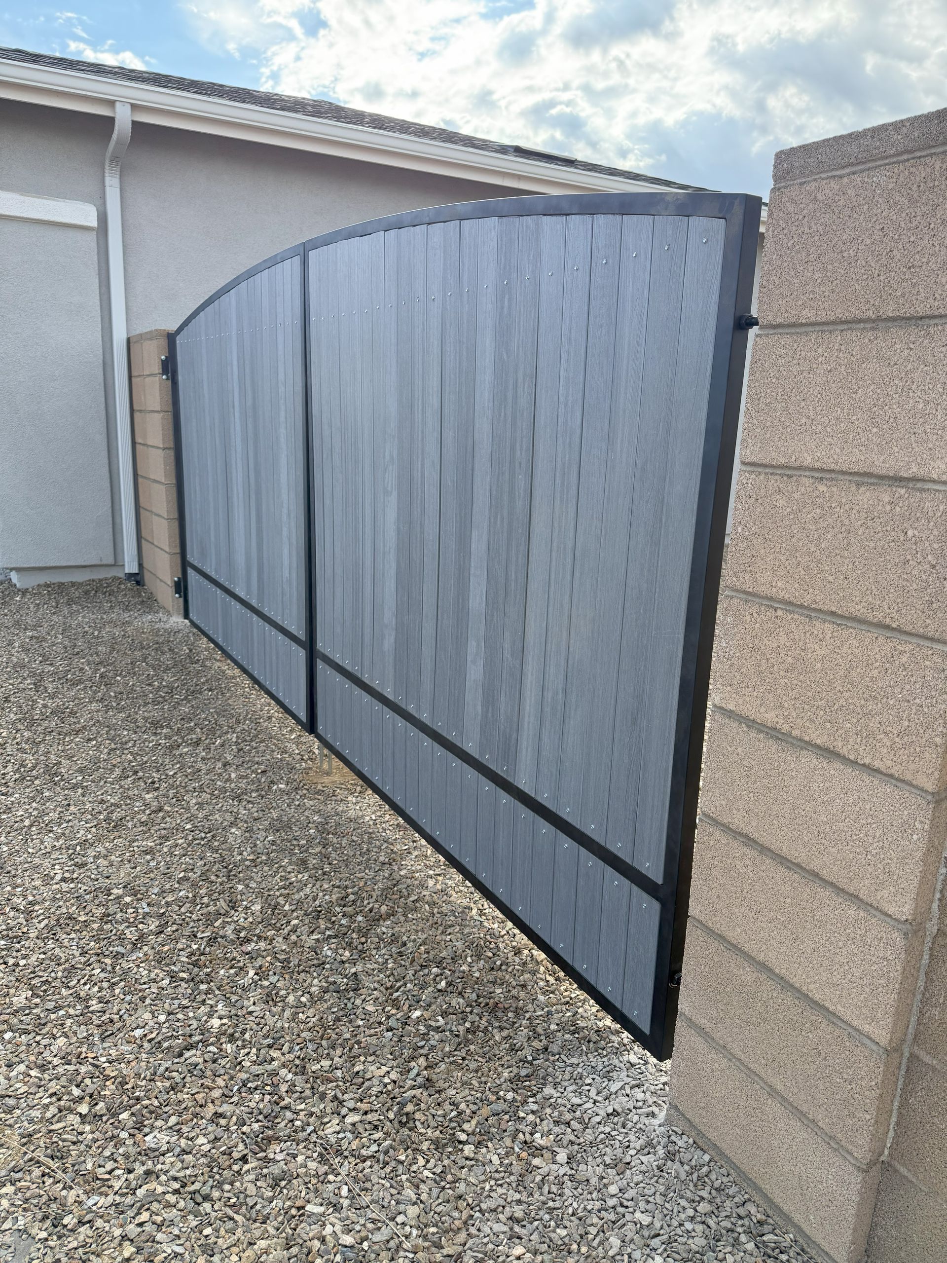 Gray, arched-top driveway gate framed in black, flanked by brick and gravel.