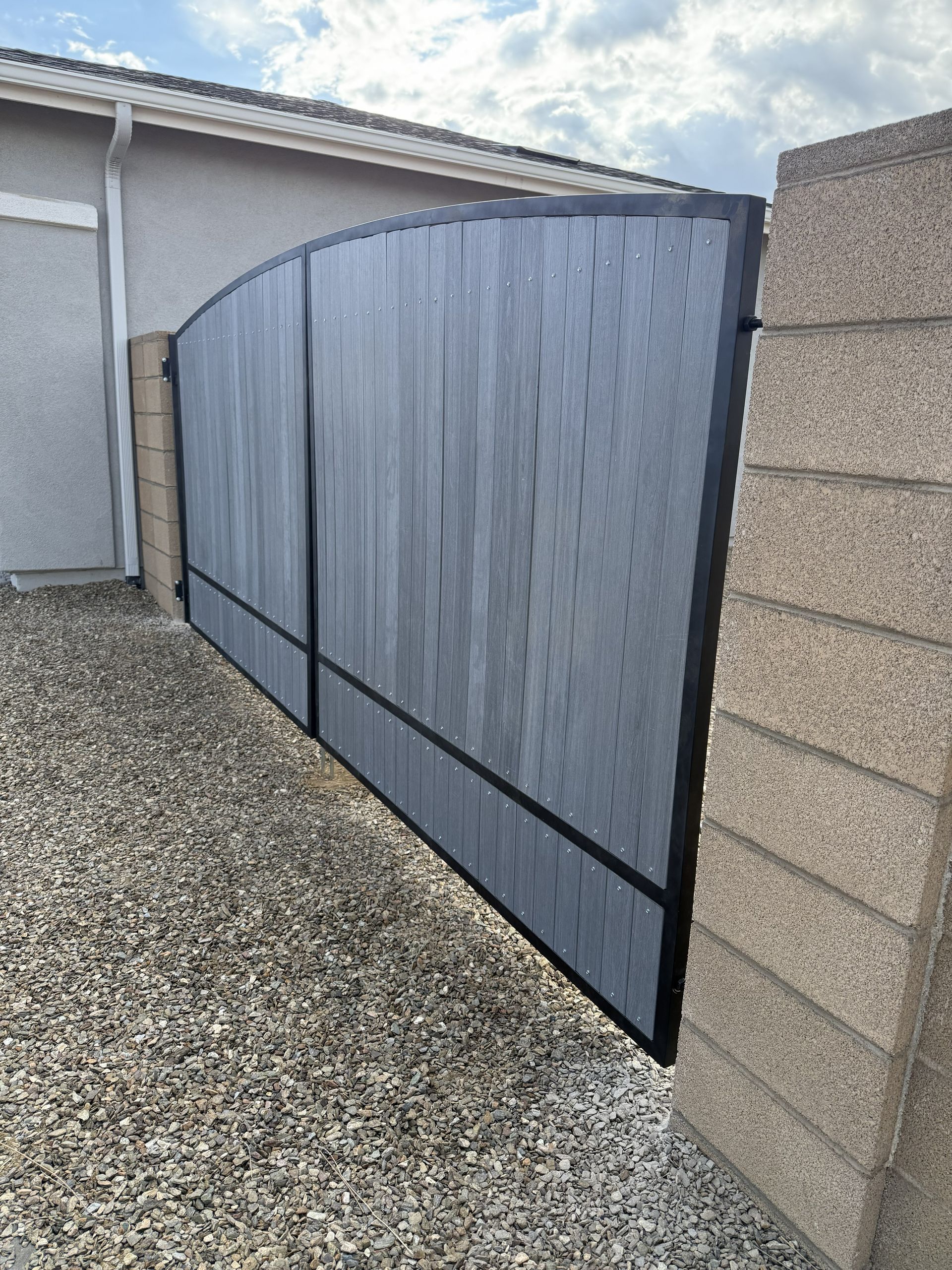 A grey, curved gate with a black metal frame installed between two concrete block walls over a gravel ground.