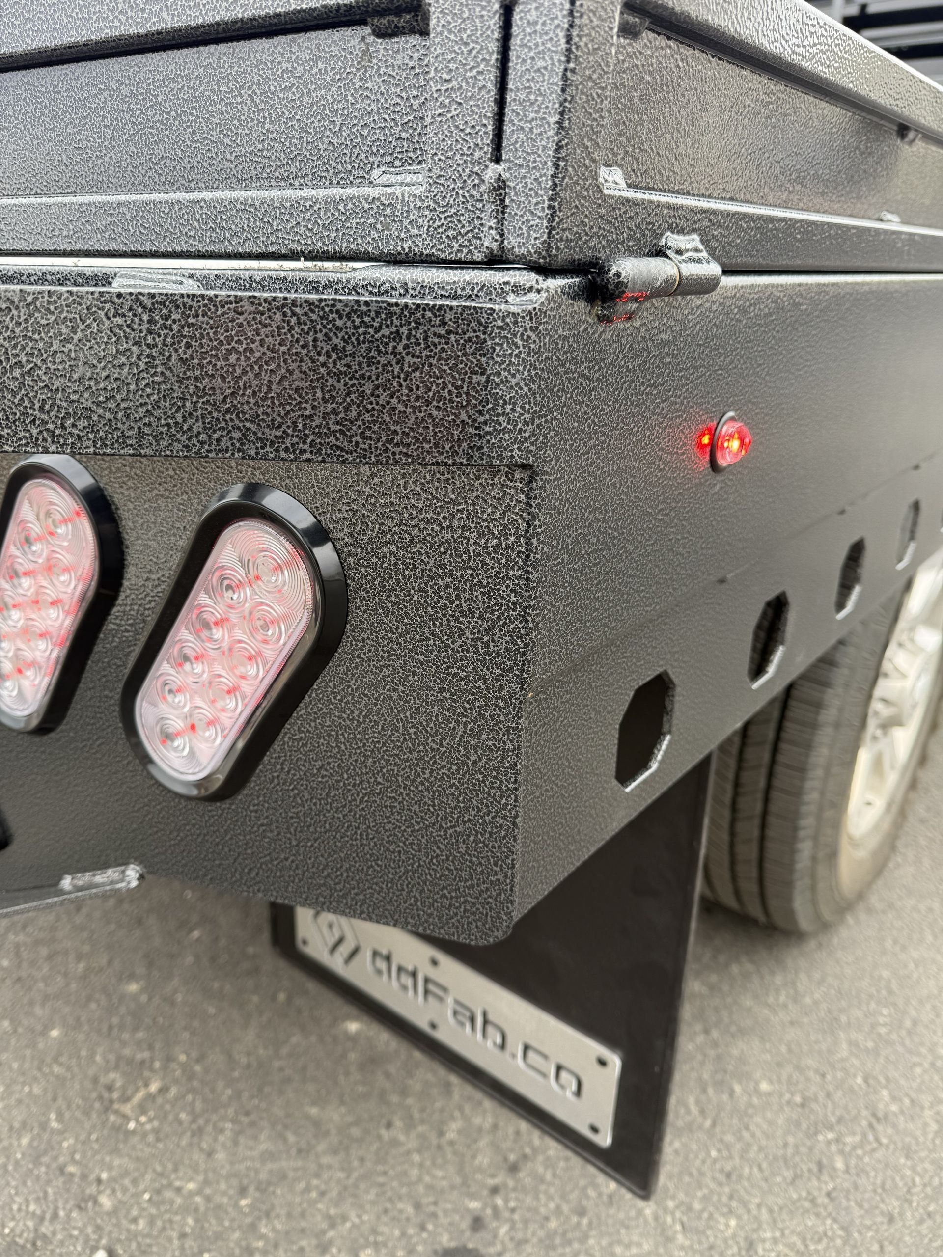The rear corner of a flatbed truck with a textured black finish, featuring two oval tail lights and a mud flap with branding.