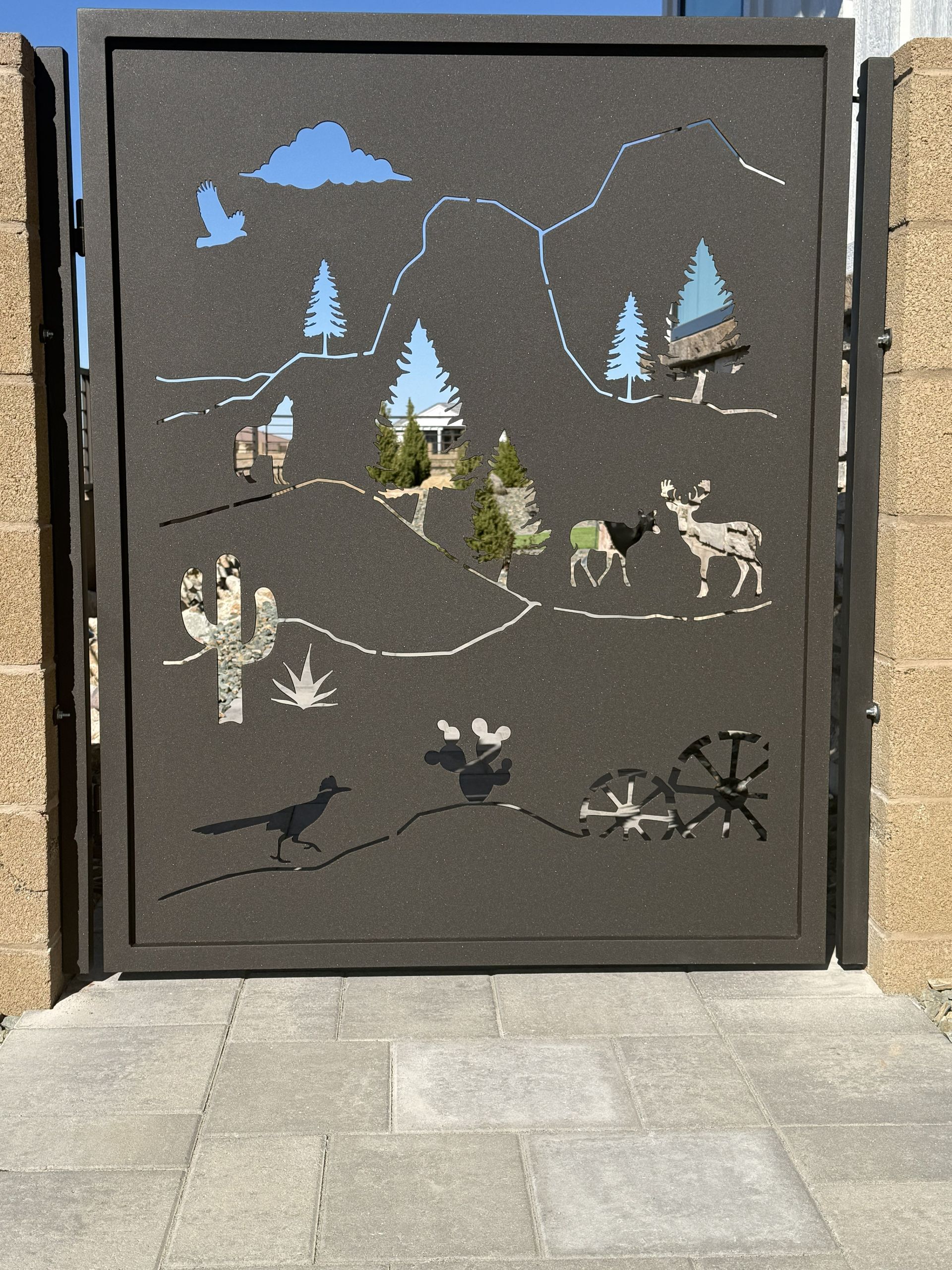 A dark metal gate with laser-cut desert scenery, including cacti, animals, and mountain shapes, set between stone pillars.