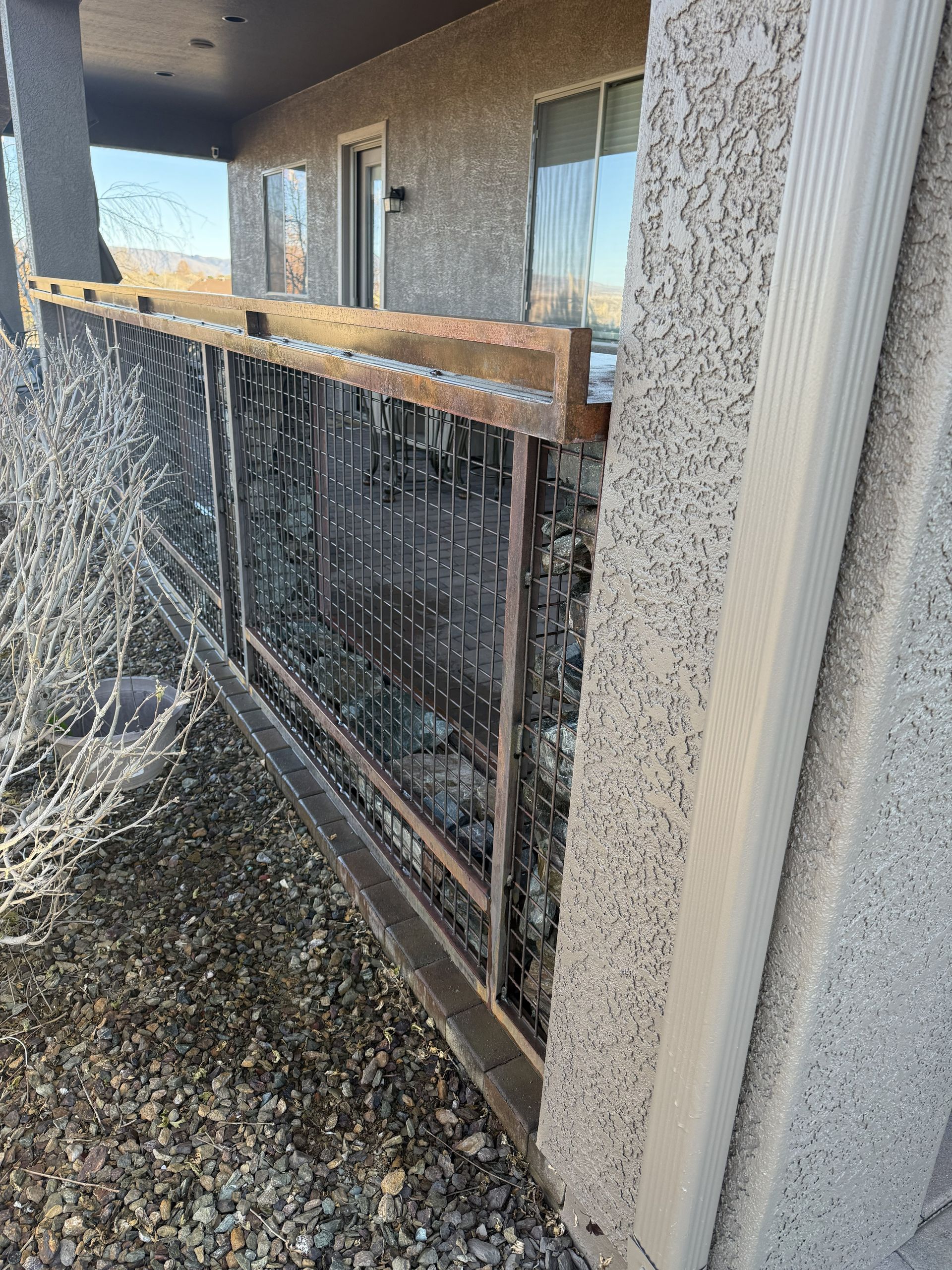 A metal privacy screen with a mesh pattern and wooden top rail, installed against a textured stucco wall on a patio.