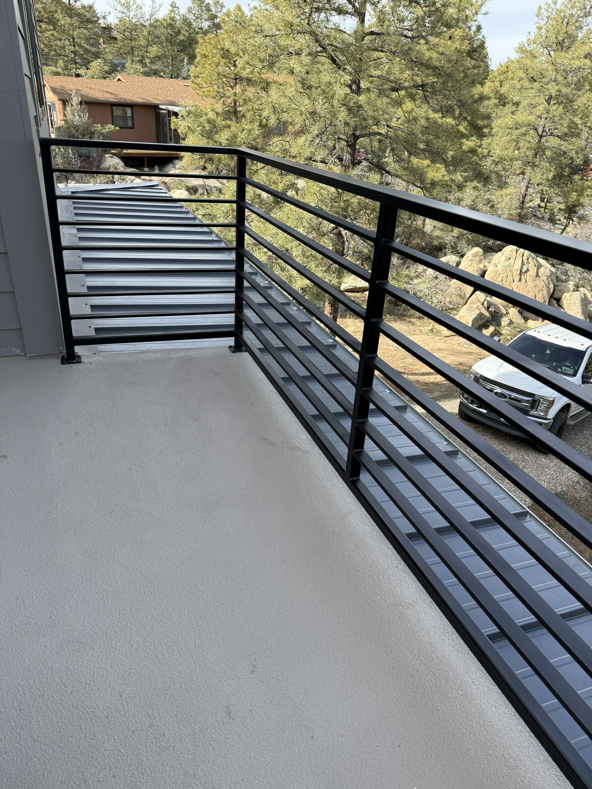 A black horizontal-rail balcony railing overlooks a wooded area and a parked vehicle.