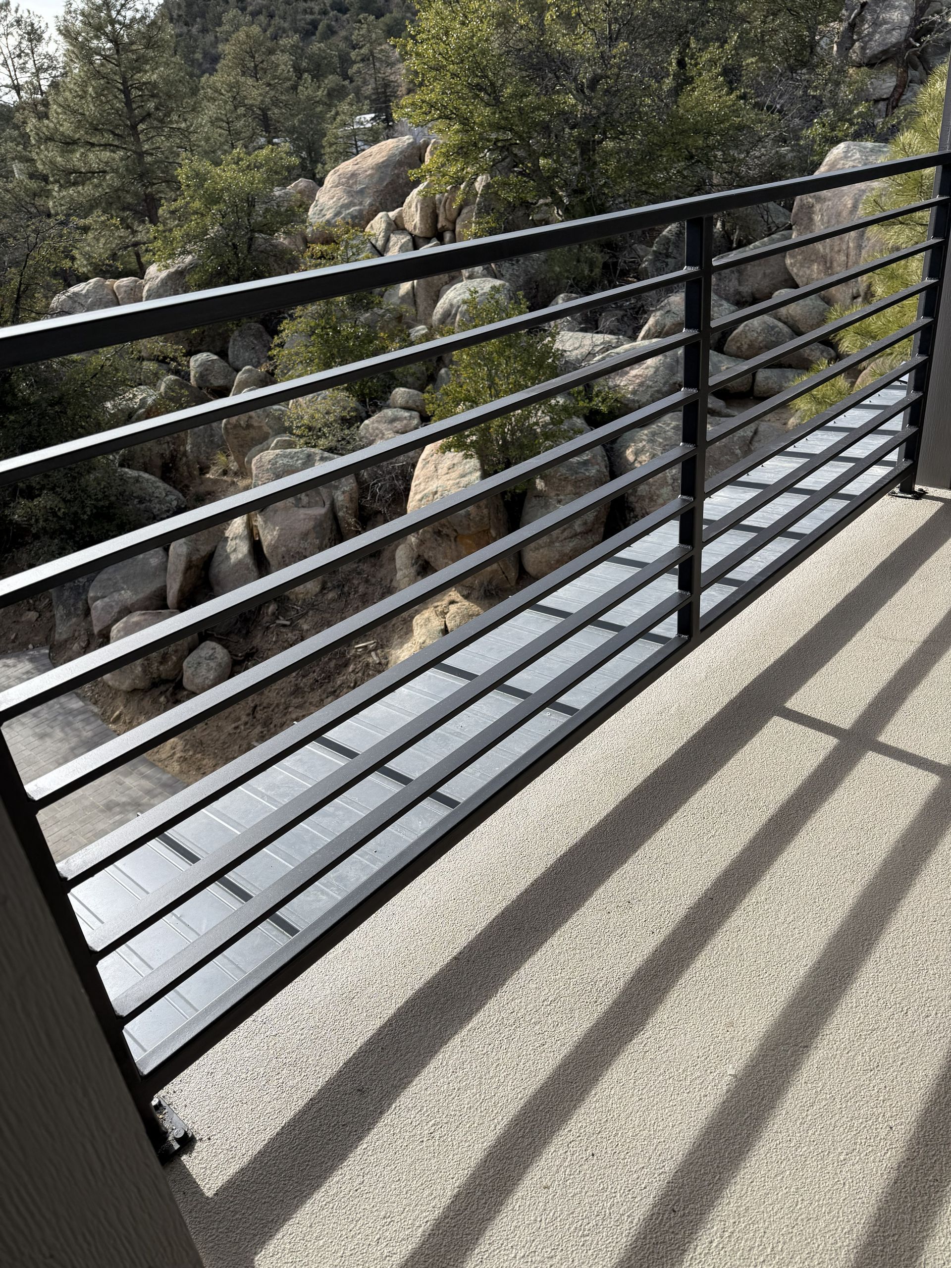 A black metal horizontal bar railing on a balcony overlooking a rocky, wooded hillside in daylight.