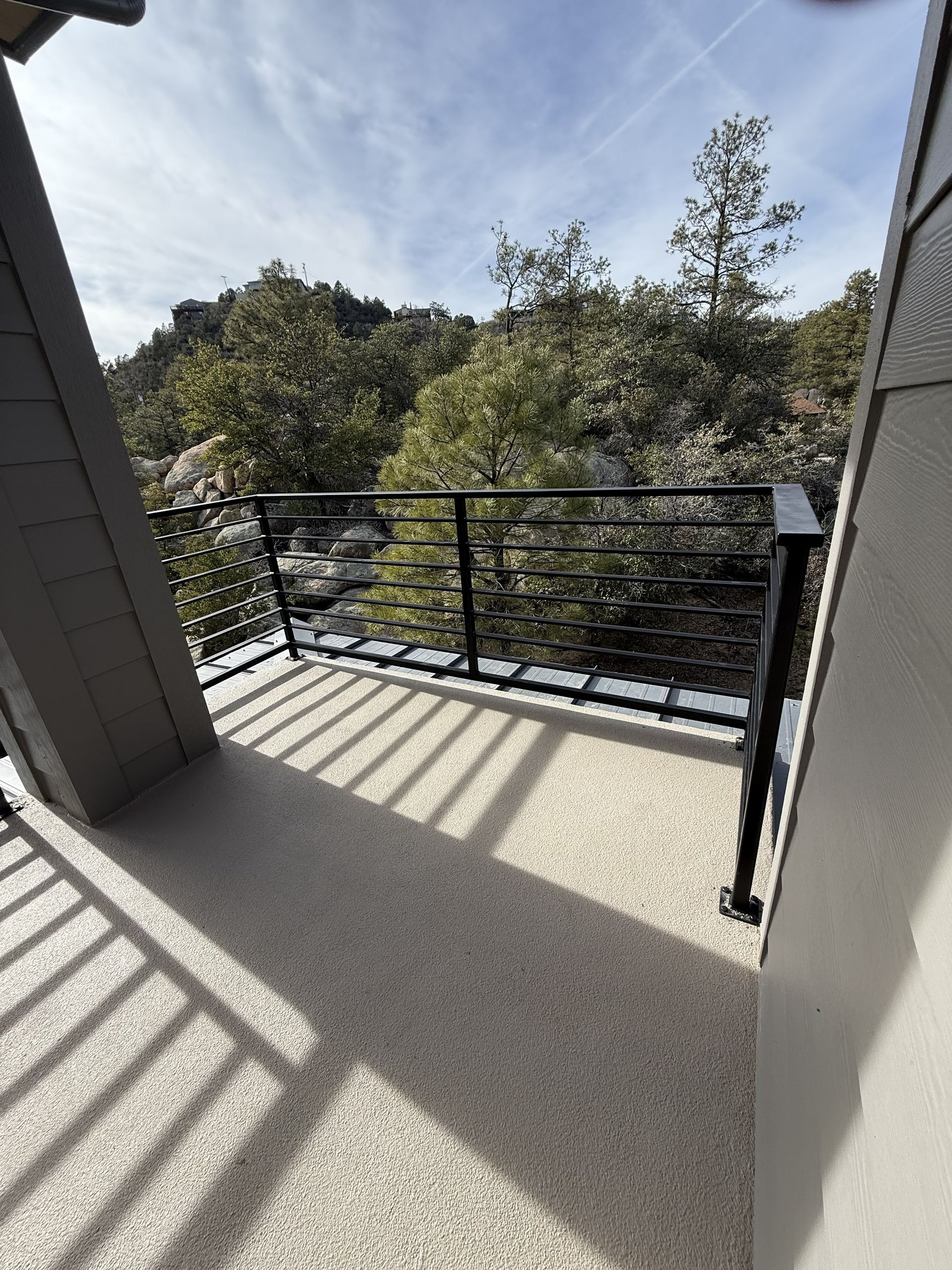 A balcony with a speckled beige floor and a black metal railing overlooking trees and rocky terrain under a bright sky.