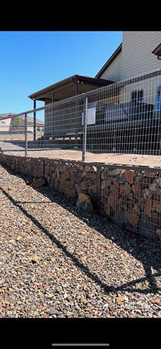 A house with a retaining wall made of rocks. A metal fence is on top. A gravel driveway is in front.