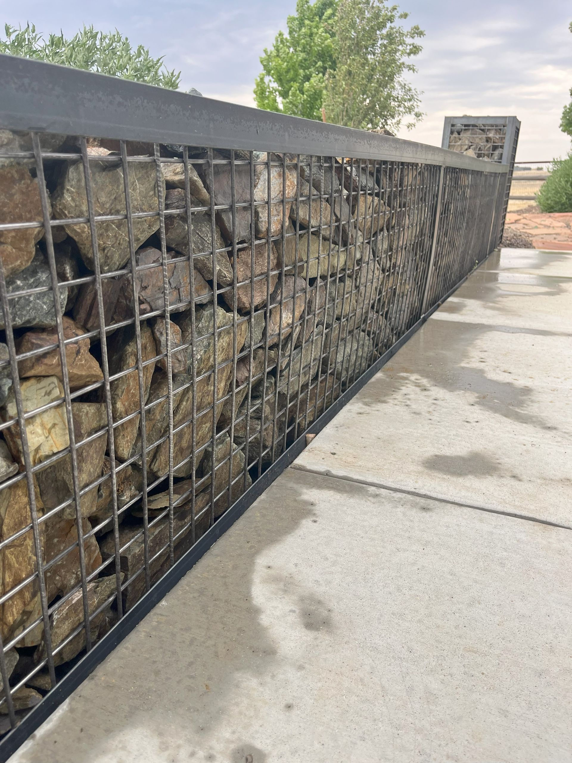 Gabion wall filled with rocks next to a concrete walkway. Black metal frame.