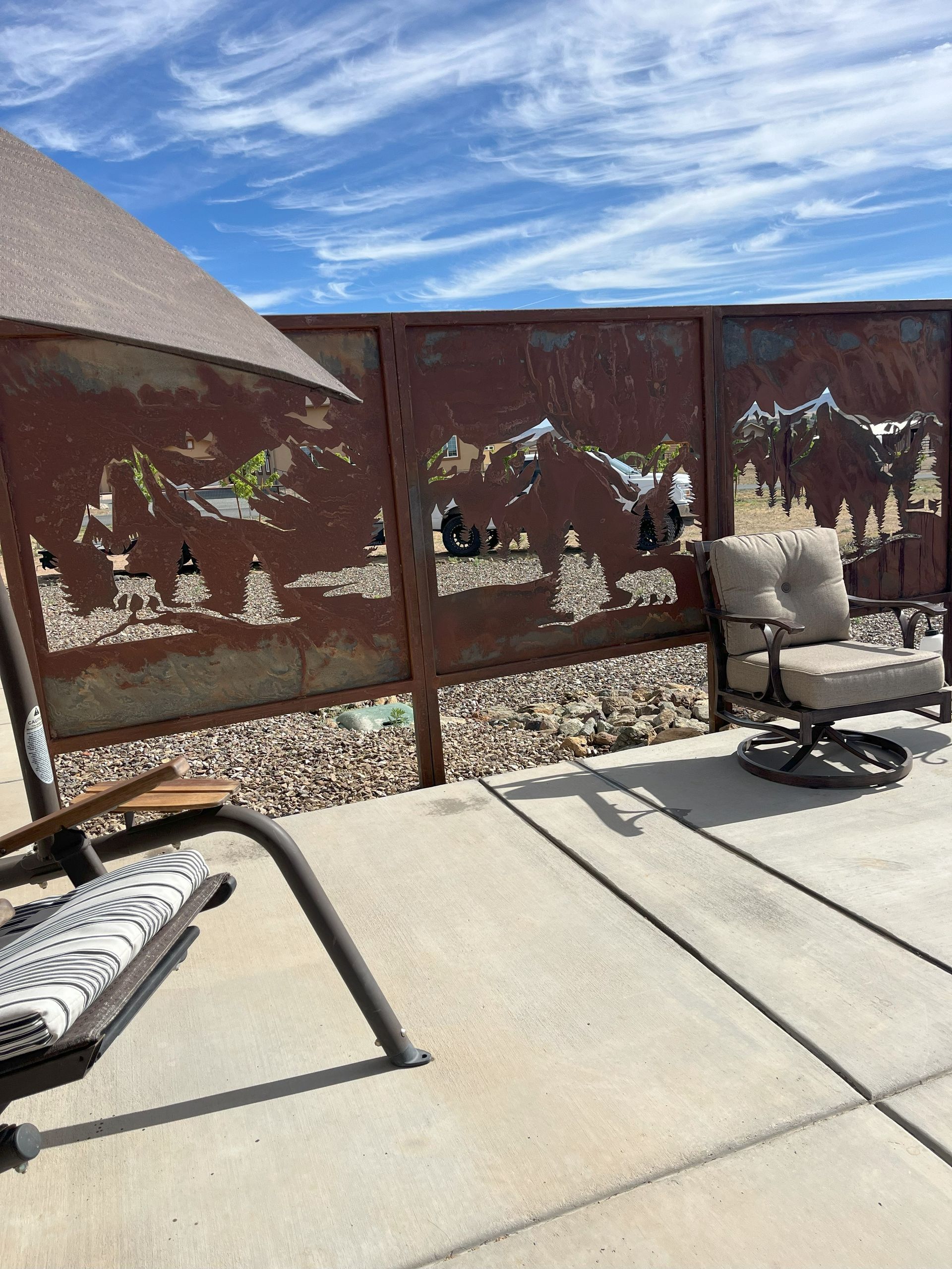 Rusty metal privacy fence with cut-out mountain scene; patio furniture on concrete patio; blue sky.