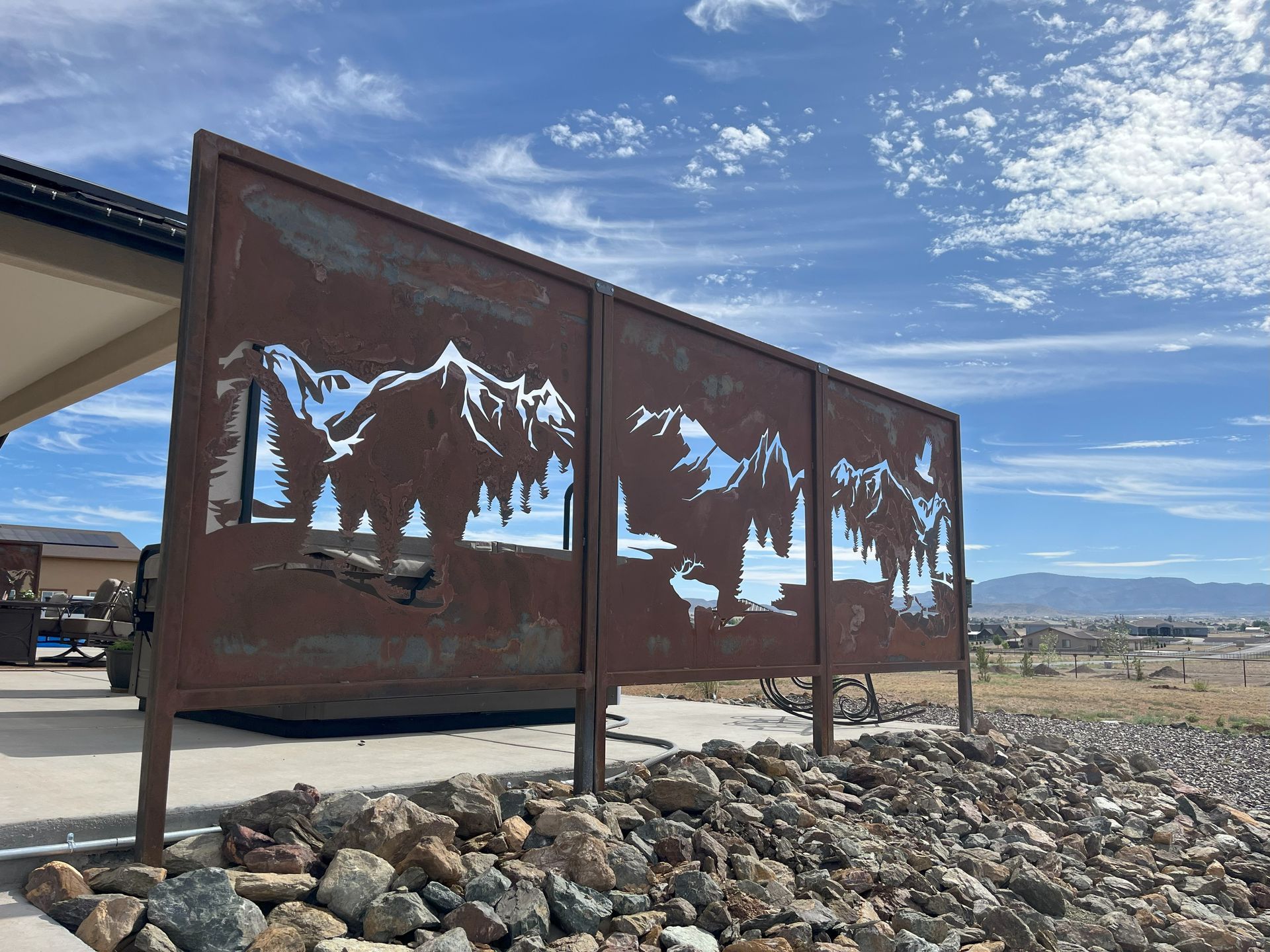 Rusty metal screen with mountain scene cutouts; near a building and rocks, under blue sky.