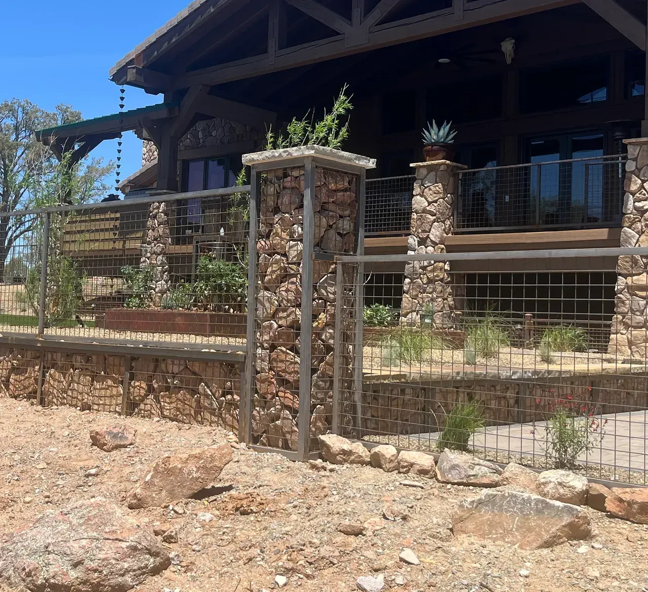 Stone and metal fence surrounding a large, rustic wooden house.
