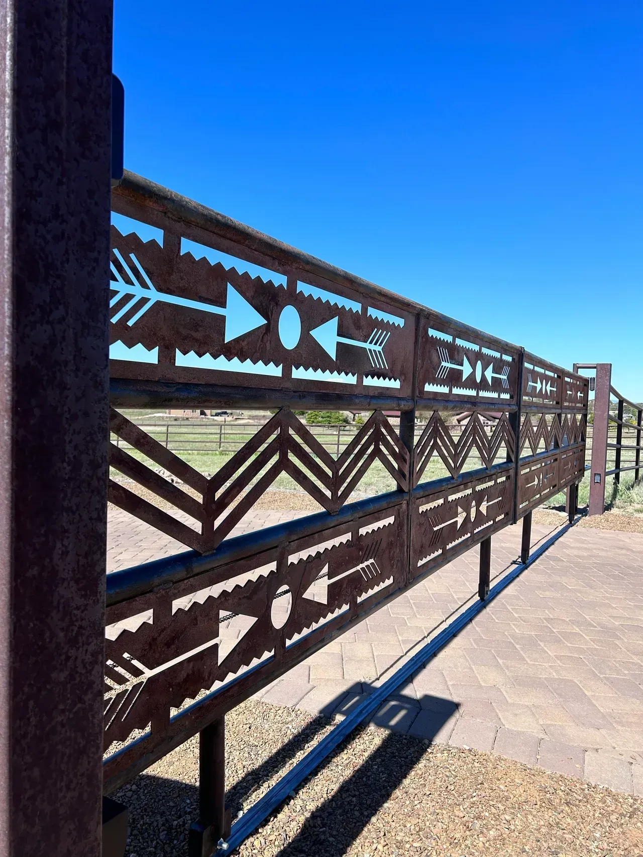 Rusty metal fence with cut-out arrow, zig-zag, and circle designs against a blue sky.