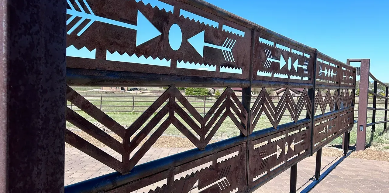 Metal fence with arrow and zigzag cutouts against a blue sky, framing a field.