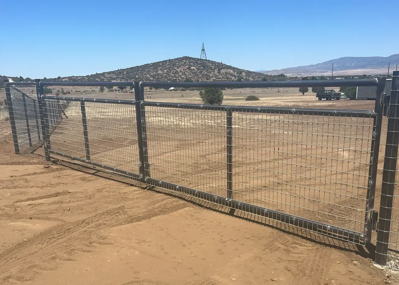 A black metal gate with wire mesh, set in a dry, sandy landscape under a clear blue sky.