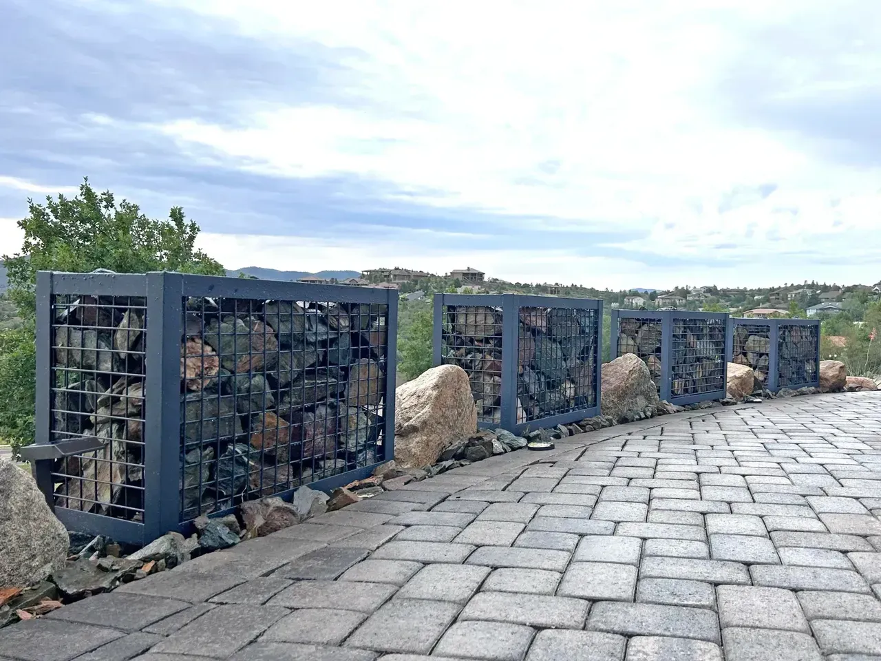 Gabion wall fence filled with rocks, along a brick pathway, cloudy sky in the background.
