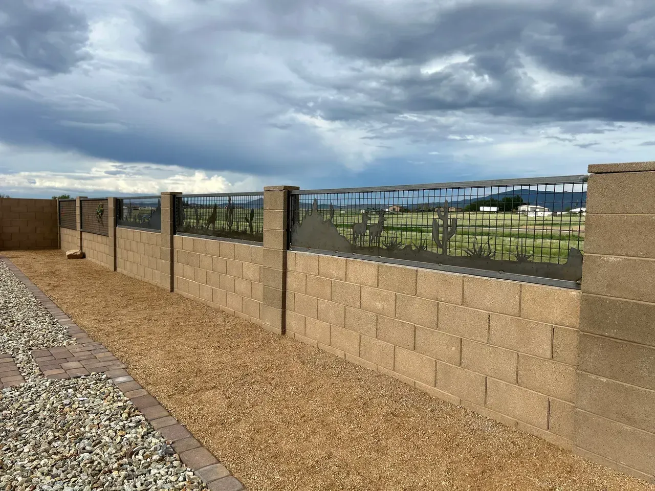 Brown block wall fence with decorative metal top, gravel path, and cloudy sky.