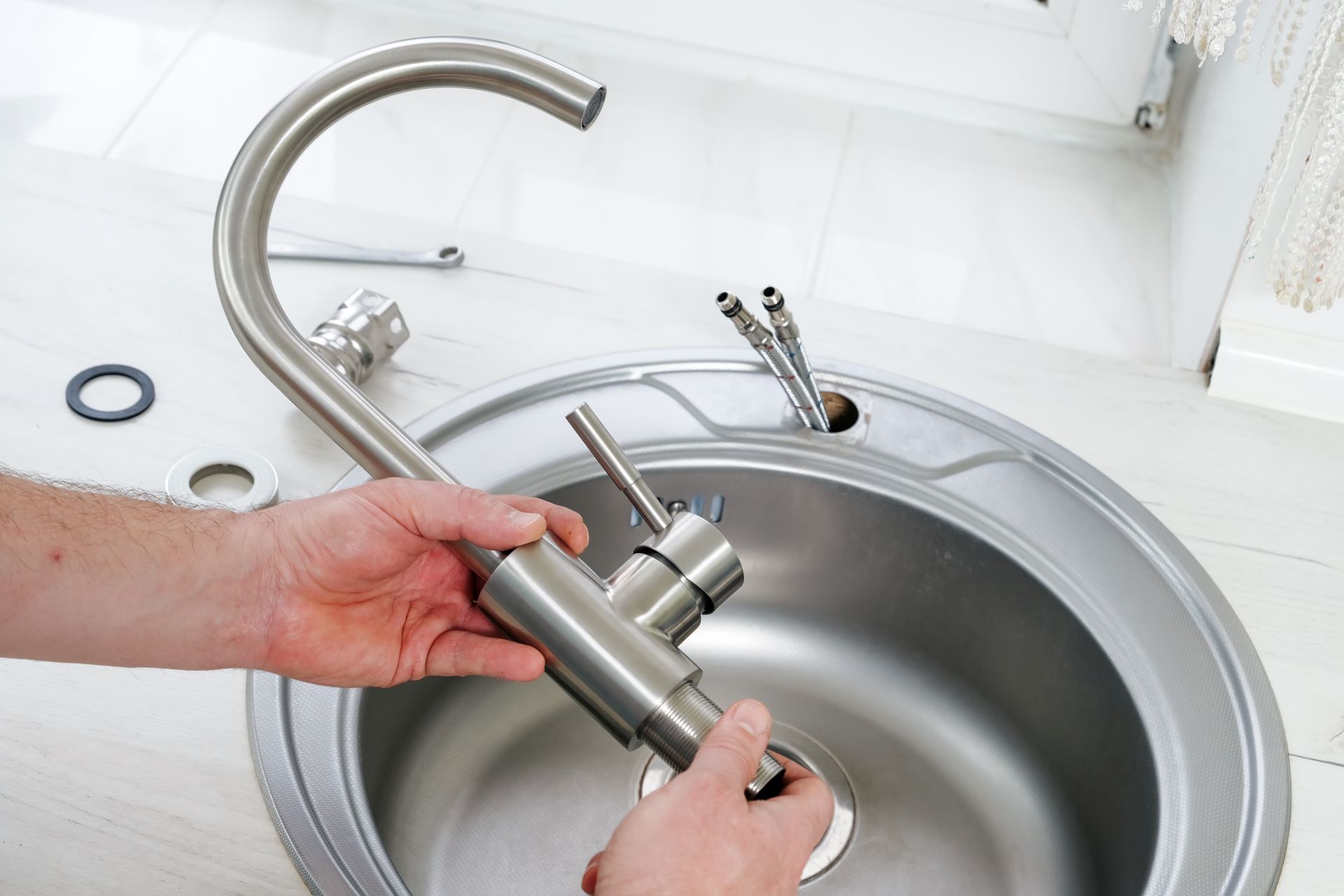 Hands removing a stainless-steel faucet from a sink, with tools on a white countertop.