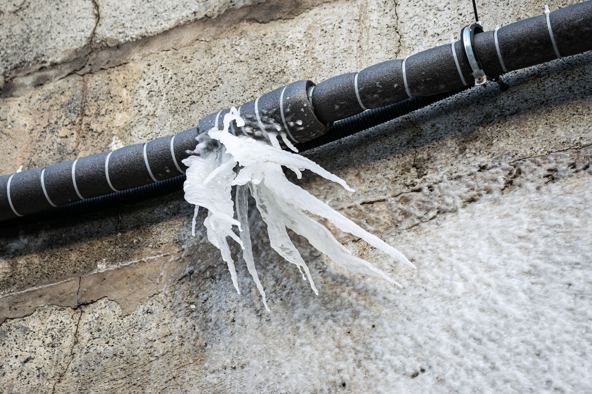 White icicle hanging from a black pipe on a concrete wall