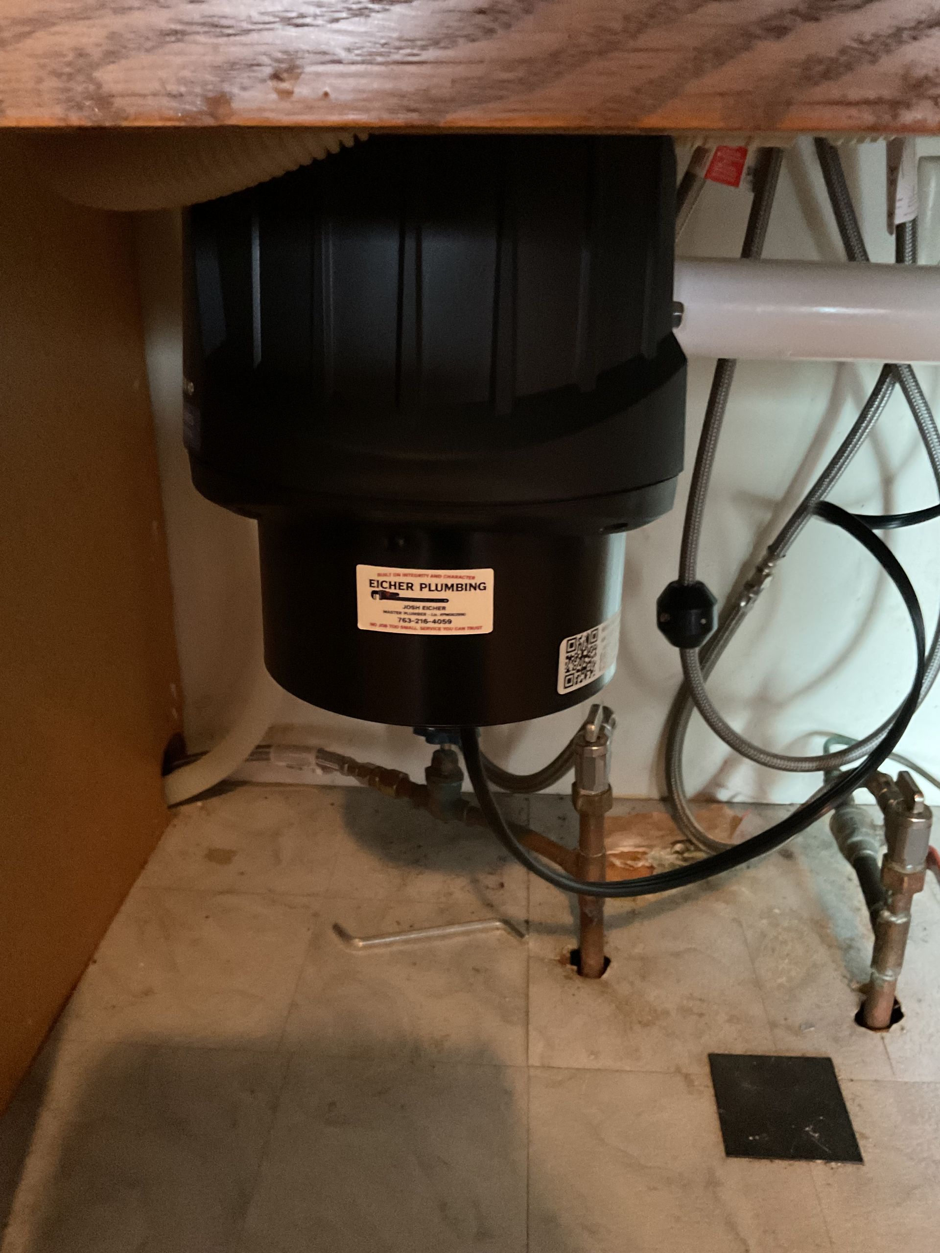 Black trash can under a sink with exposed pipes and cords in a utility area