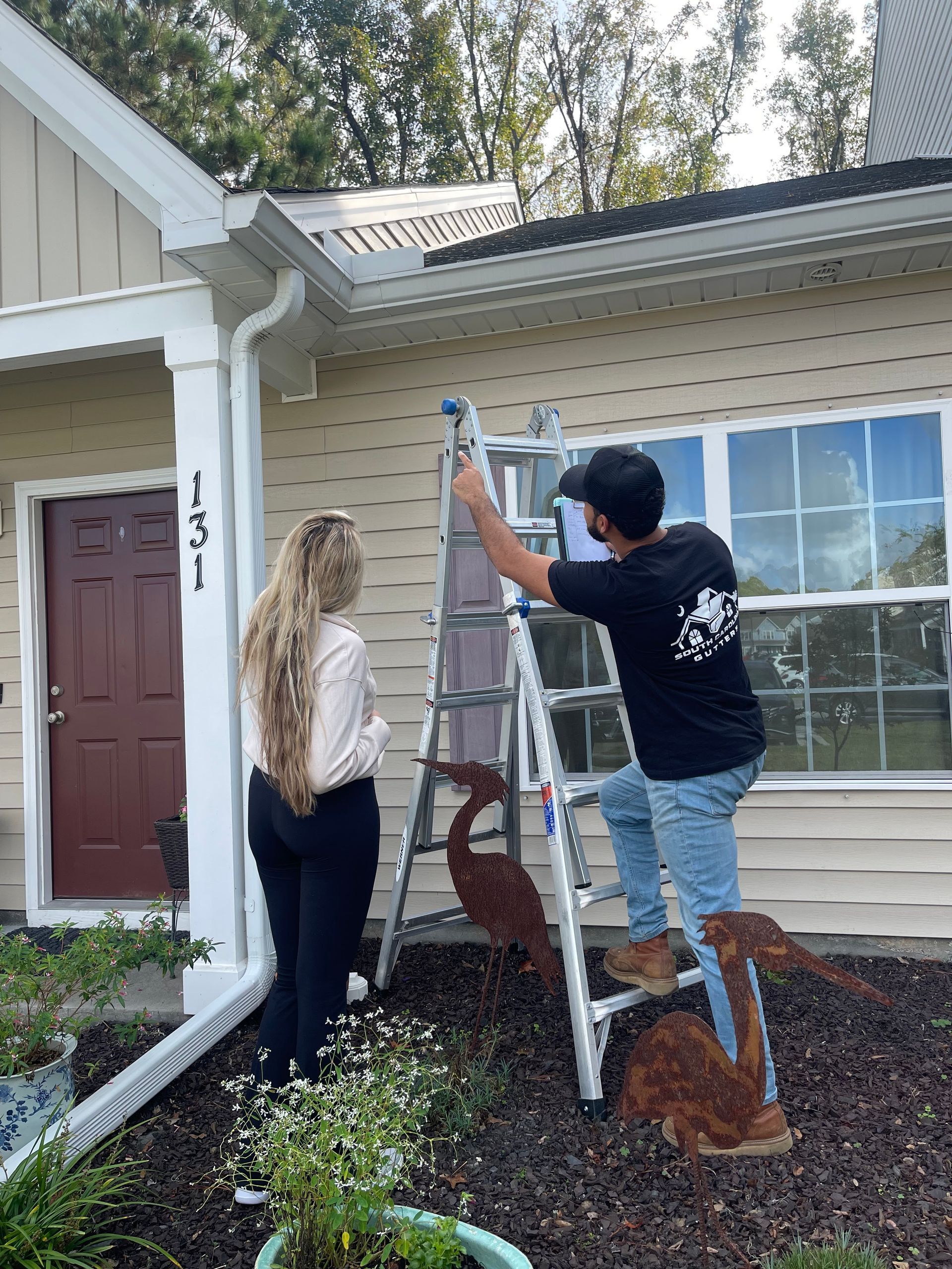 A person on a ladder paints the exterior trim of a house while another person stands nearby, observing the work.
