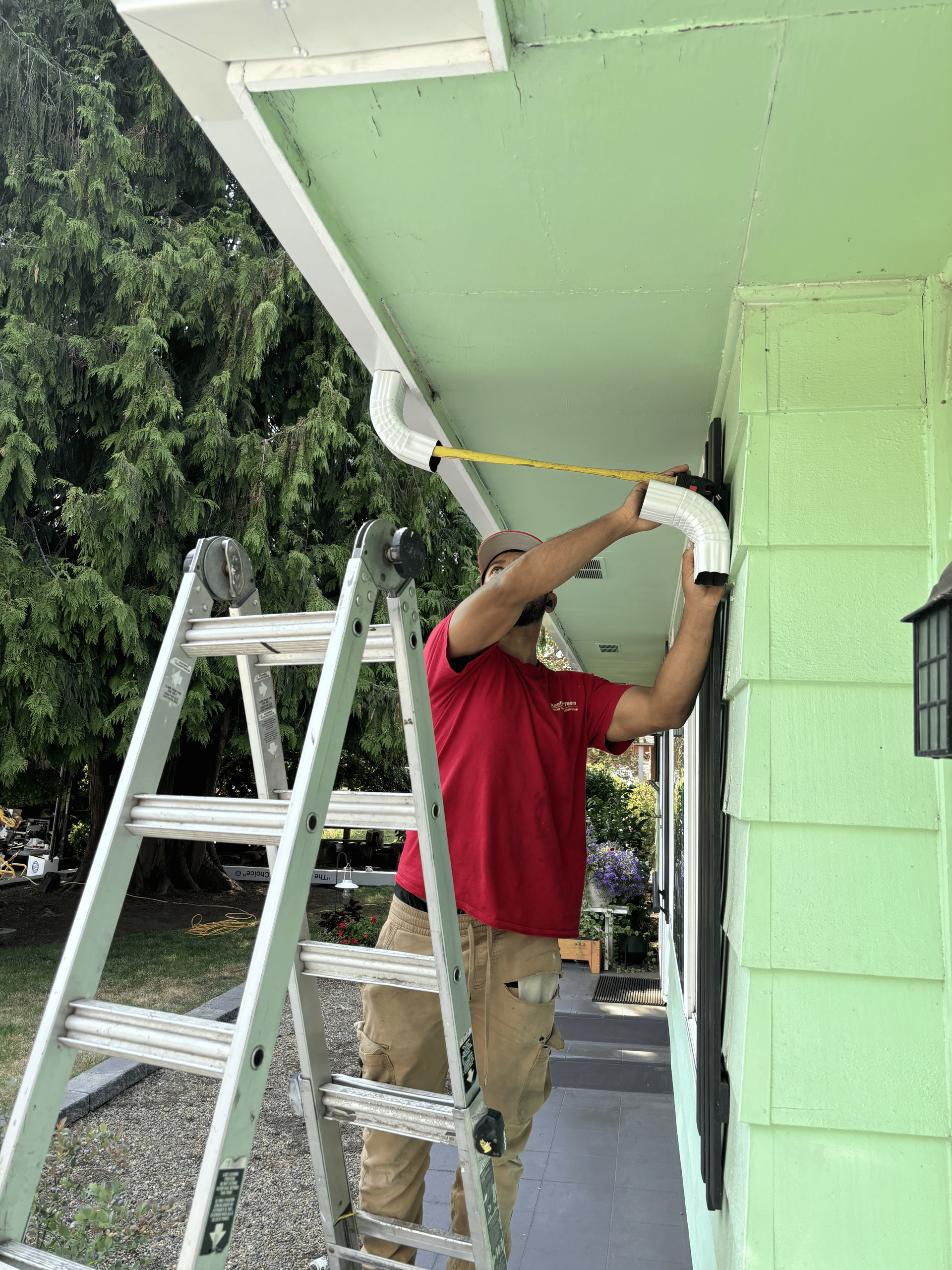 A person wearing a red shirt and tan pants stands on a ladder, using a tape measure to install white gutters on a house.