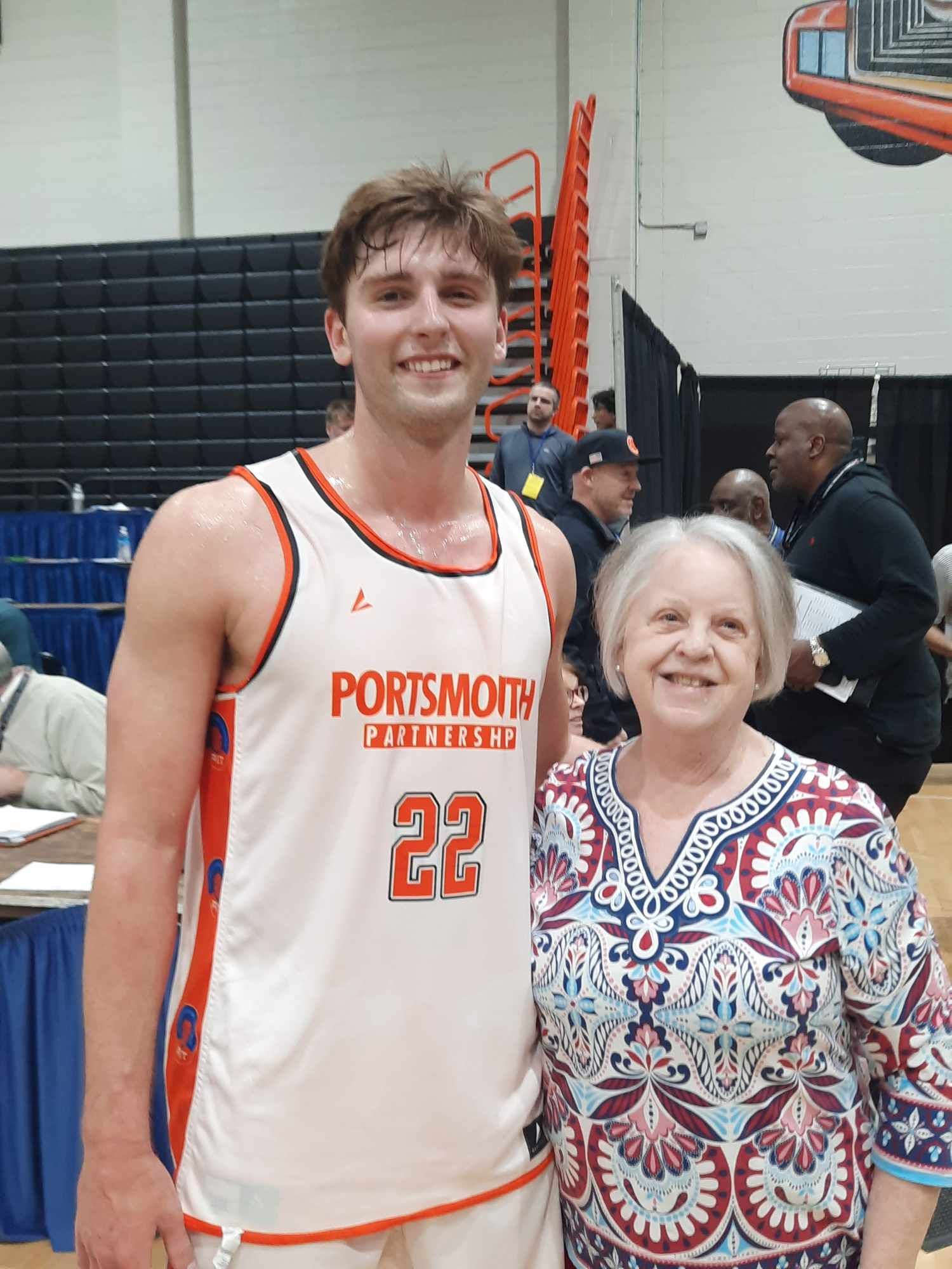 Basketball player in white Portsmouth jersey stands smiling beside an older woman indoors.