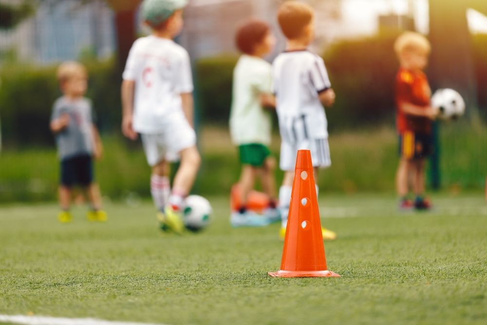 Orange traffic cone on green field, children in soccer uniforms, playing.
