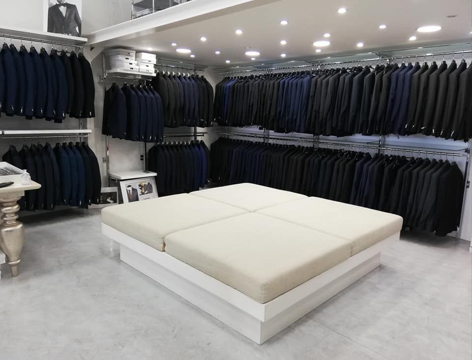 Negozio di abbigliamento moderno con abiti scuri appesi a grucce e un letto beige esposto al centro.