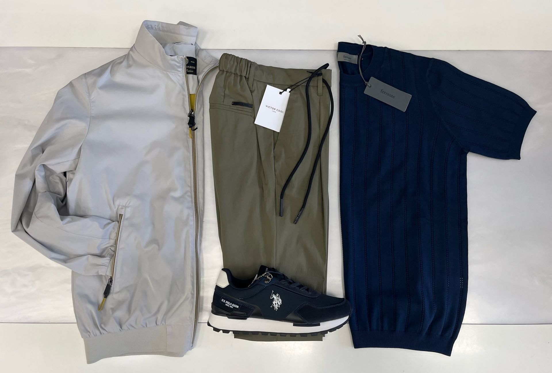 Giacca grigia, pantaloni verde oliva, camicia blu scuro e scarpe da ginnastica nere disposti su uno sfondo bianco.