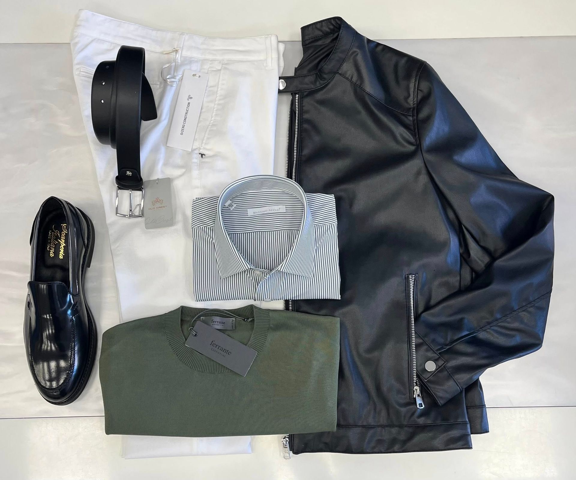 Su una superficie bianca sono disposti diversi capi di abbigliamento e accessori da uomo, tra cui una giacca nera, scarpe, cintura e camicie.