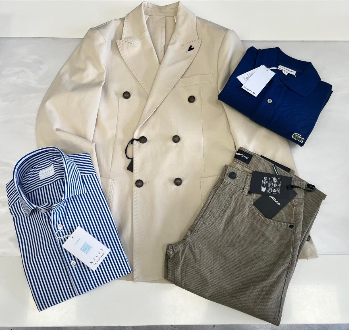Vari capi di abbigliamento piegati: blazer color crema, camicia a righe blu, polo blu navy e pantaloni grigi su sfondo bianco.