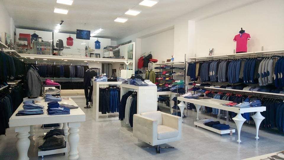 Interno di un negozio di abbigliamento con scaffali pieni di jeans, camicie piegate e tavoli espositivi bianchi.