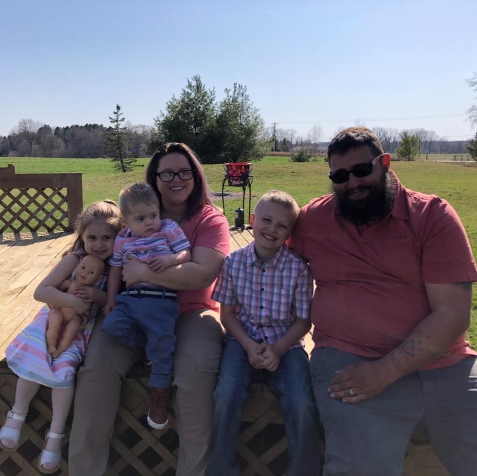 Family sits on a wooden deck outdoors; sunny day. Woman holds toddler; man has his arm around another child.