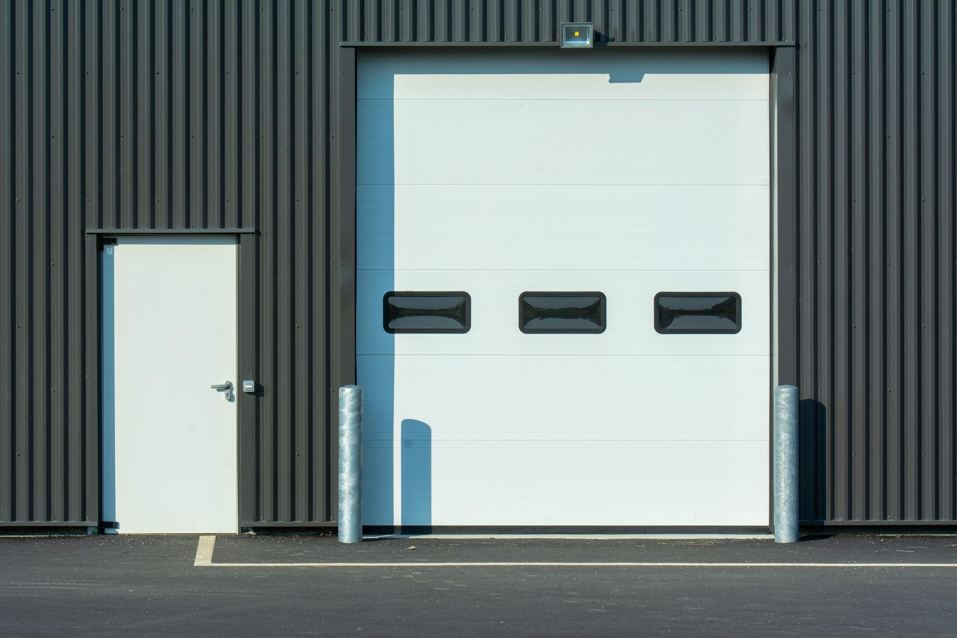 White overhead door and side door in a gray building. Black asphalt and silver posts.