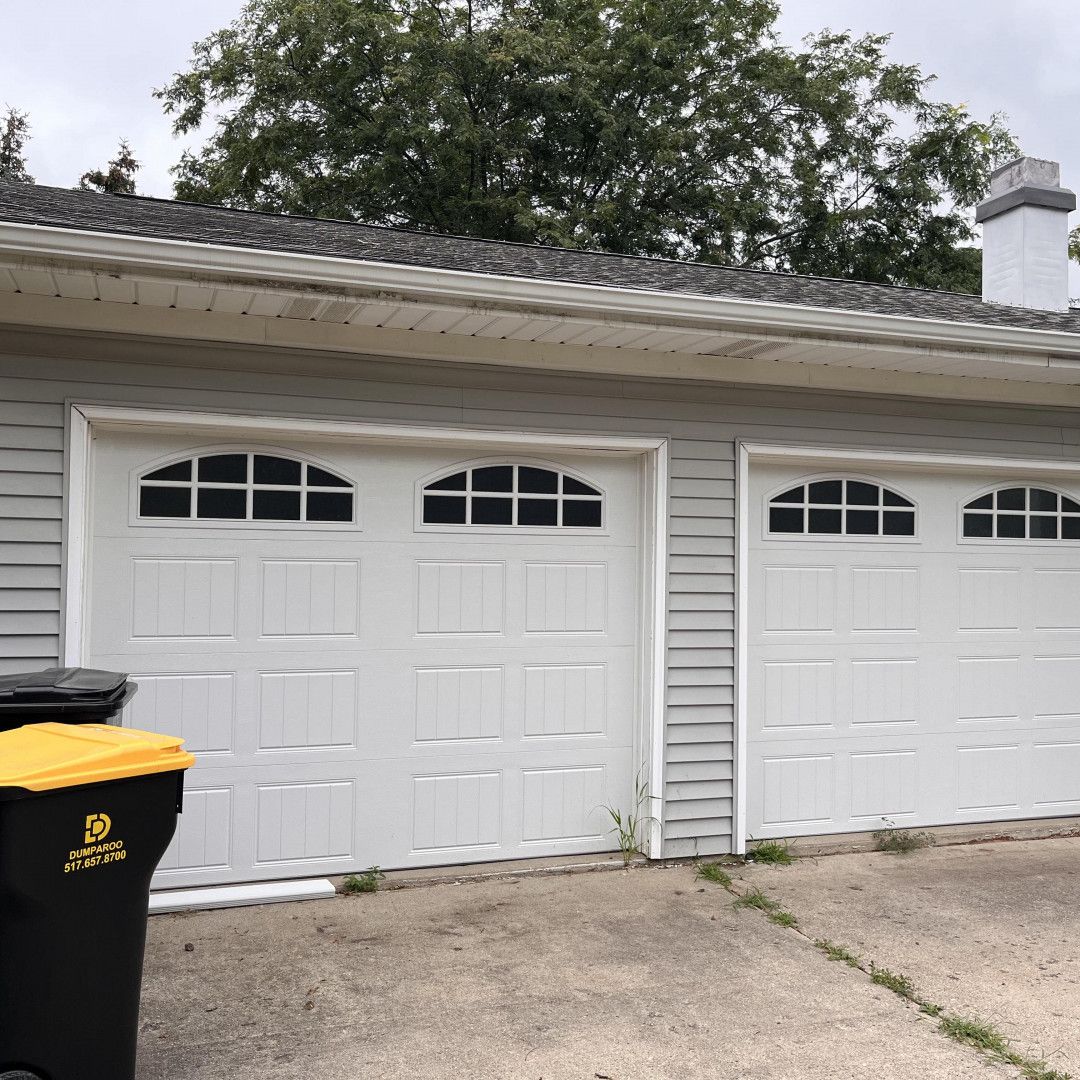 Two white garage doors with arched window panels. A black trash can is in front of the left door. Gray siding, concrete, and trees.