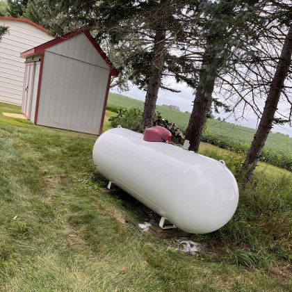 White propane tank on grass near a shed and trees. Red cap on top.