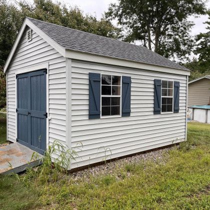 White shed with blue door and shutters, two windows, gray roof, grassy yard.