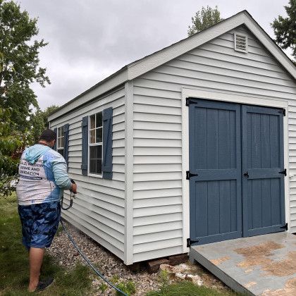 Person spraying water on a light gray shed with blue doors and shutters, standing outside on a cloudy day.