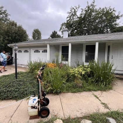A person near a pressure washer on a sidewalk in front of a house with overgrown landscaping and a cloudy sky.