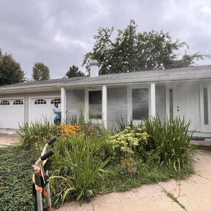 Person pressure washing a gray house, white garage doors, and front door, overgrown garden in front.