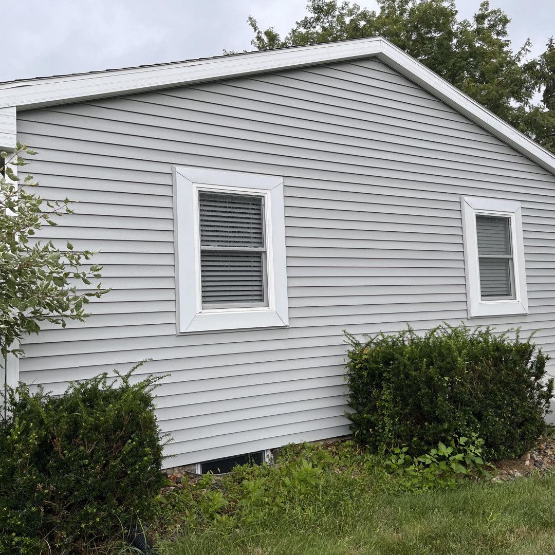 Light gray house siding with white trim around windows and roofline; two windows, bushes in front.