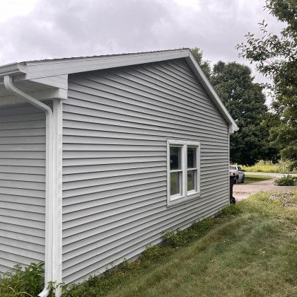 Gray house with white trim, window, and gutter. Green grass and trees in background. Cloudy sky.