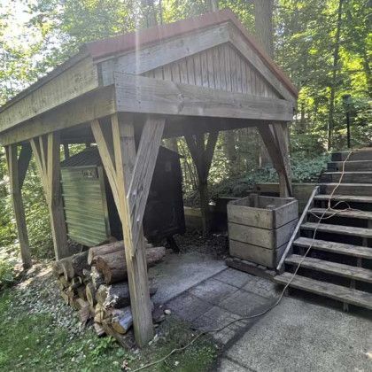 Wooden shelter with firewood, storage, and stairs in a wooded area.
