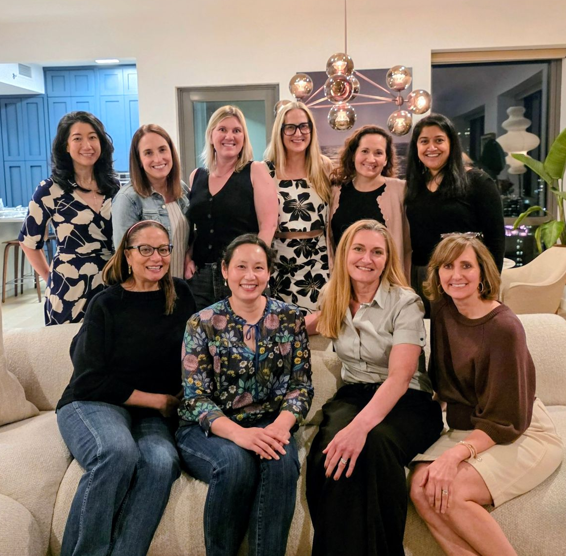 Group of women posing together in a bright living room, smiling in coordinated black, white, and beige outfits.